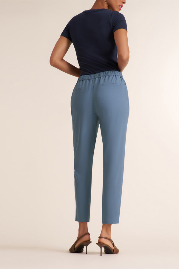 A model shown from the back wearing slate blue cropped trousers, a navy blue short-sleeve top, and brown strappy heels against a solid beige studio background. | Exclude | Grid Hover
