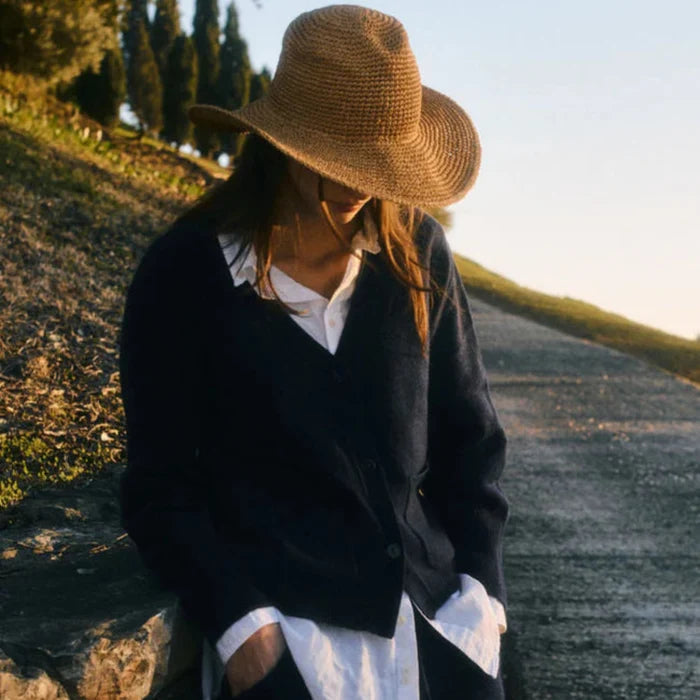 Person wearing a straw hat and dark jacket standing by a stone wall with a scenic background.