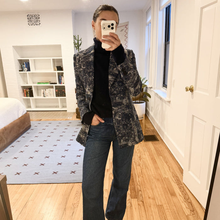 Person taking a mirror selfie wearing a patterned coat, black shirt, and jeans in a bedroom.
