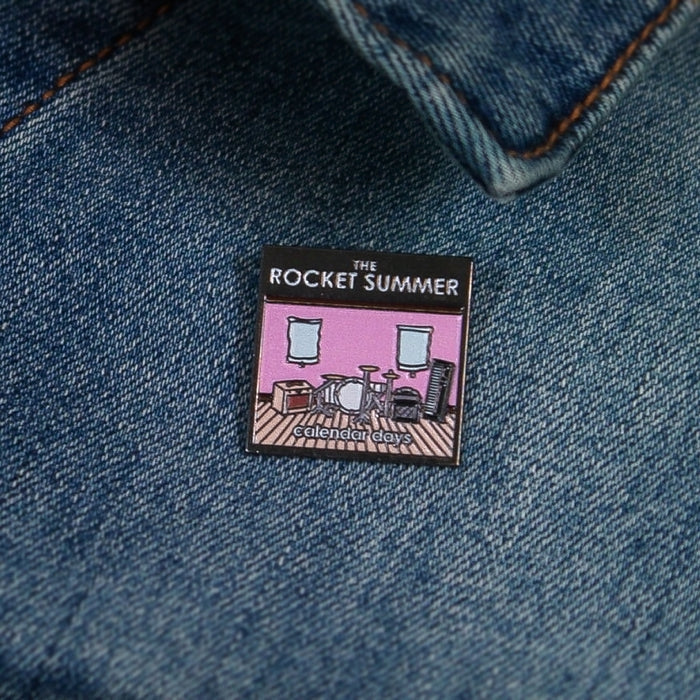 A 1 x 1 enamel pin by The Rocket Summer features their name above a pink room with musical instruments and calendar dates at the bottom, all set on denim.