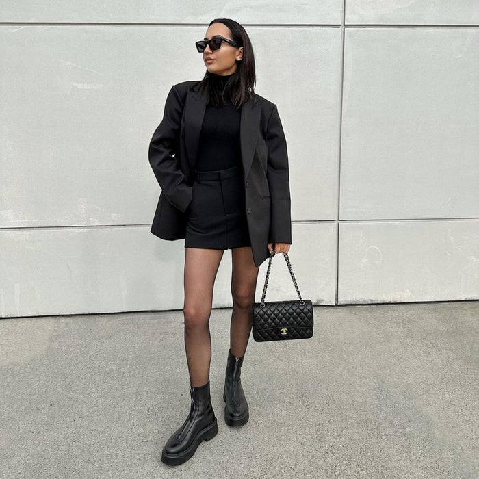 Person wearing a black outfit with a blazer, skirt, and boots, holding a black handbag against a gray wall. | Exclude