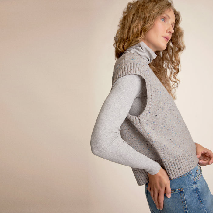 Person wearing a gray sweater vest and blue jeans on a beige background