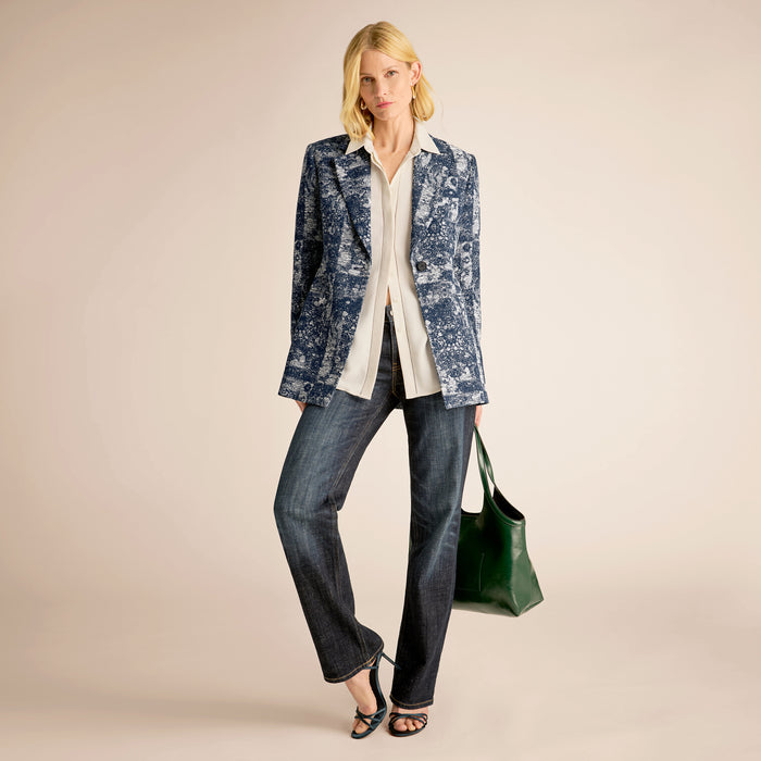 Woman wearing a blue patterned jacket, white shirt, and jeans, holding a green bag on a beige background