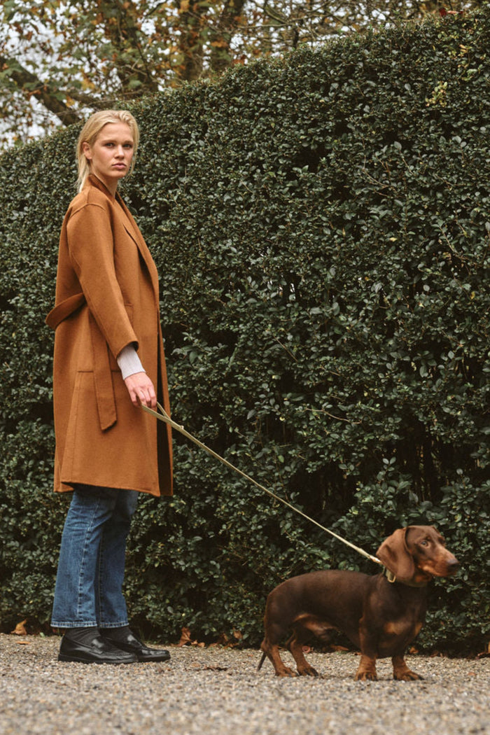 Woman in a brown coat walking a dachshund in an outdoor setting with greenery. | Grid Hover | Exclude
