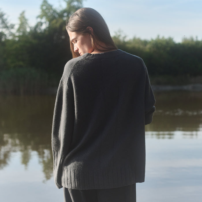 Person wearing a dark sweater standing by a body of water with trees in the background