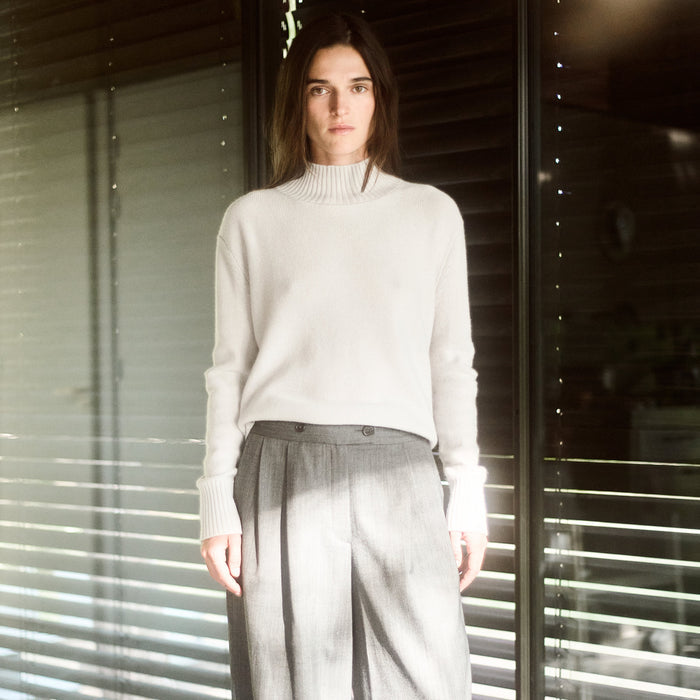 Person wearing a light-colored sweater and gray pants standing in front of a window with blinds.