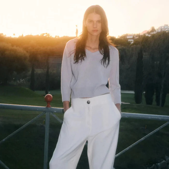 Person wearing a light gray top and white pants standing outdoors with a sunset background.
