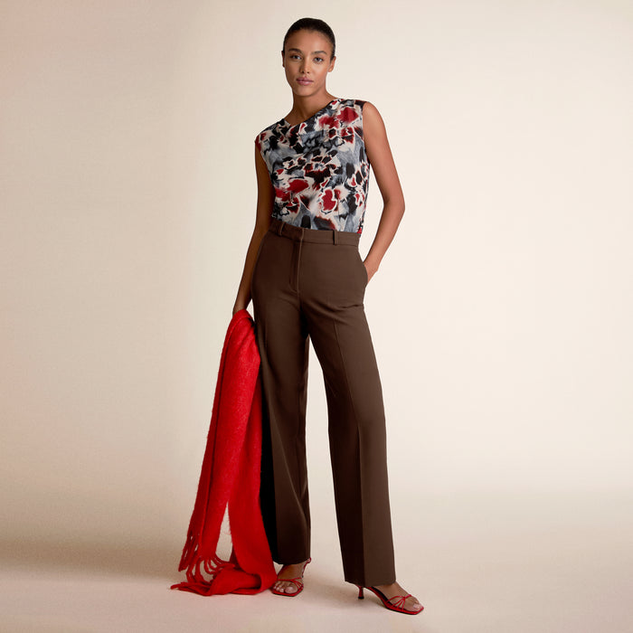 Woman wearing a floral top and brown pants with a red scarf, standing against a beige background | Lead