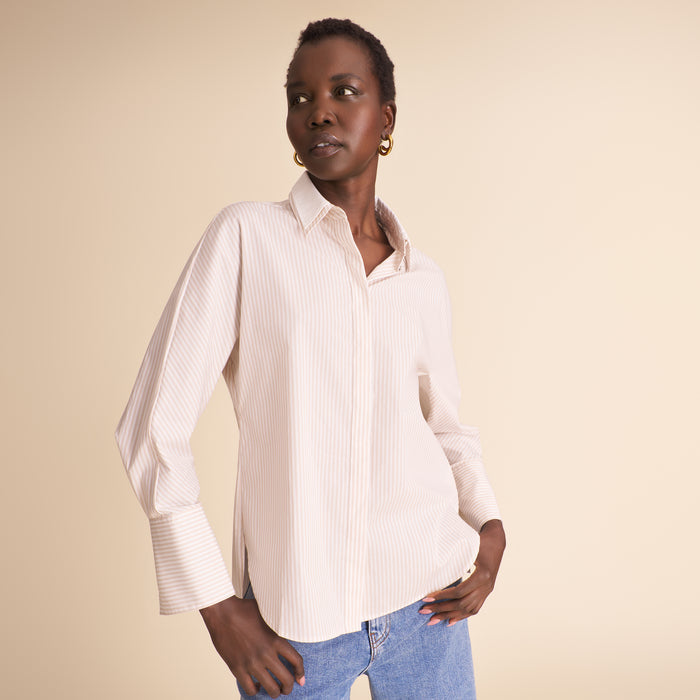 A woman modeling a light beige and white vertically striped button-down shirt with blue jeans, set against a solid beige studio background. | Lead