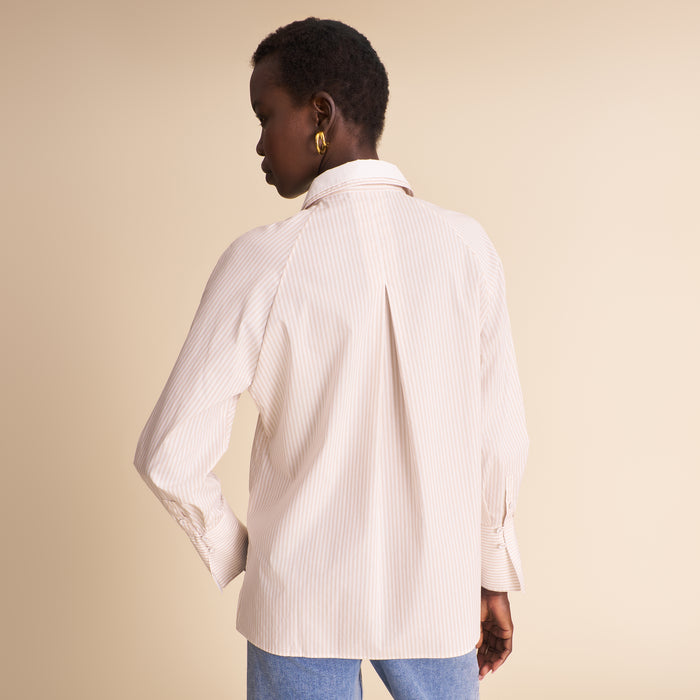 A woman shown from the back wearing a cream-colored, vertically striped button-down shirt with a wide collar and blue jeans, set against a solid beige studio background.