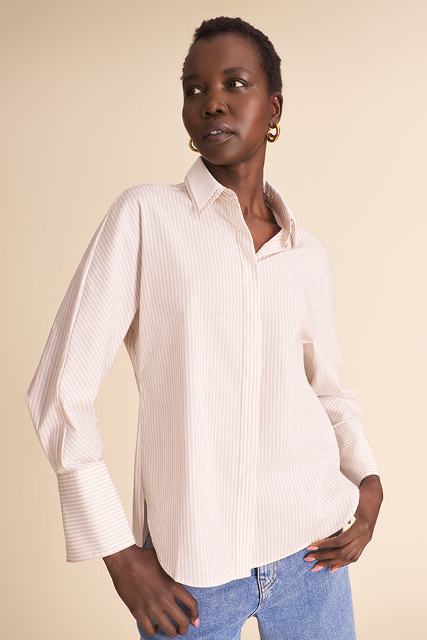 A woman wearing a light beige and white vertically striped button-down shirt with blue jeans, standing against a solid beige background. | Exclude | Grid