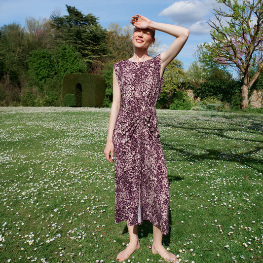 Woman in a purple floral dress standing in a garden with green grass and white flowers. | Lead
